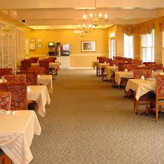 A dining room with multiple tables covered in white tablecloths and surrounded by red patterned chairs. The room is well-lit with chandeliers hanging from the ceiling and large windows with curtains allowing natural light to enter. There is a beverage station against the far wall.