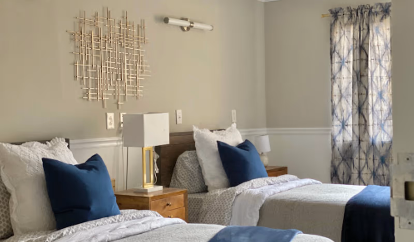 A bedroom with two twin beds, each with white and blue bedding and pillows. Between the beds is a wooden nightstand with a modern lamp. The walls are light-colored with a decorative metal wall art above the nightstand. A window with patterned blue and white curtains is visible on the right side.