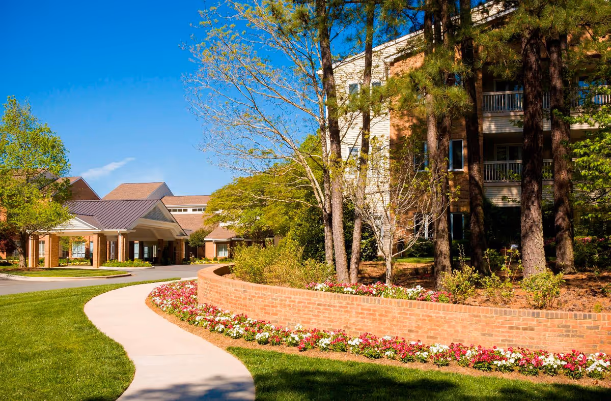 Front exterior of a brick senior living building with a covered entrance, curved sidewalk, trees, and flowerbeds.