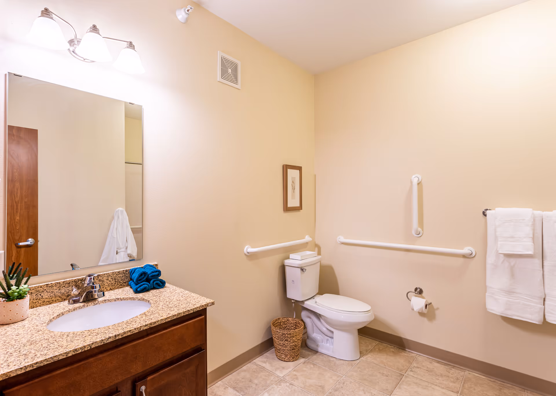 A clean and accessible bathroom with beige walls and tiled floor. It features a granite countertop with a sink, a mirror above the sink, a toilet with grab bars on the walls around it, a wicker wastebasket, a towel rack with white towels, and a small potted plant on the countertop.