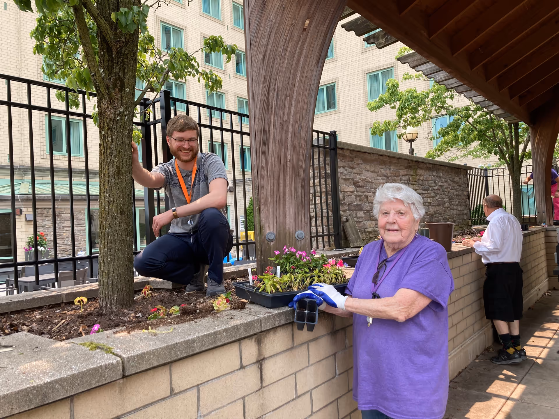 An elderly woman wearing a purple shirt and gardening gloves is planting flowers in a raised garden bed. A young man with glasses and an orange lanyard is crouching nearby, smiling and touching a tree. Another person is seen gardening in the background. The setting is an outdoor courtyard with a brick building and windows in the background.