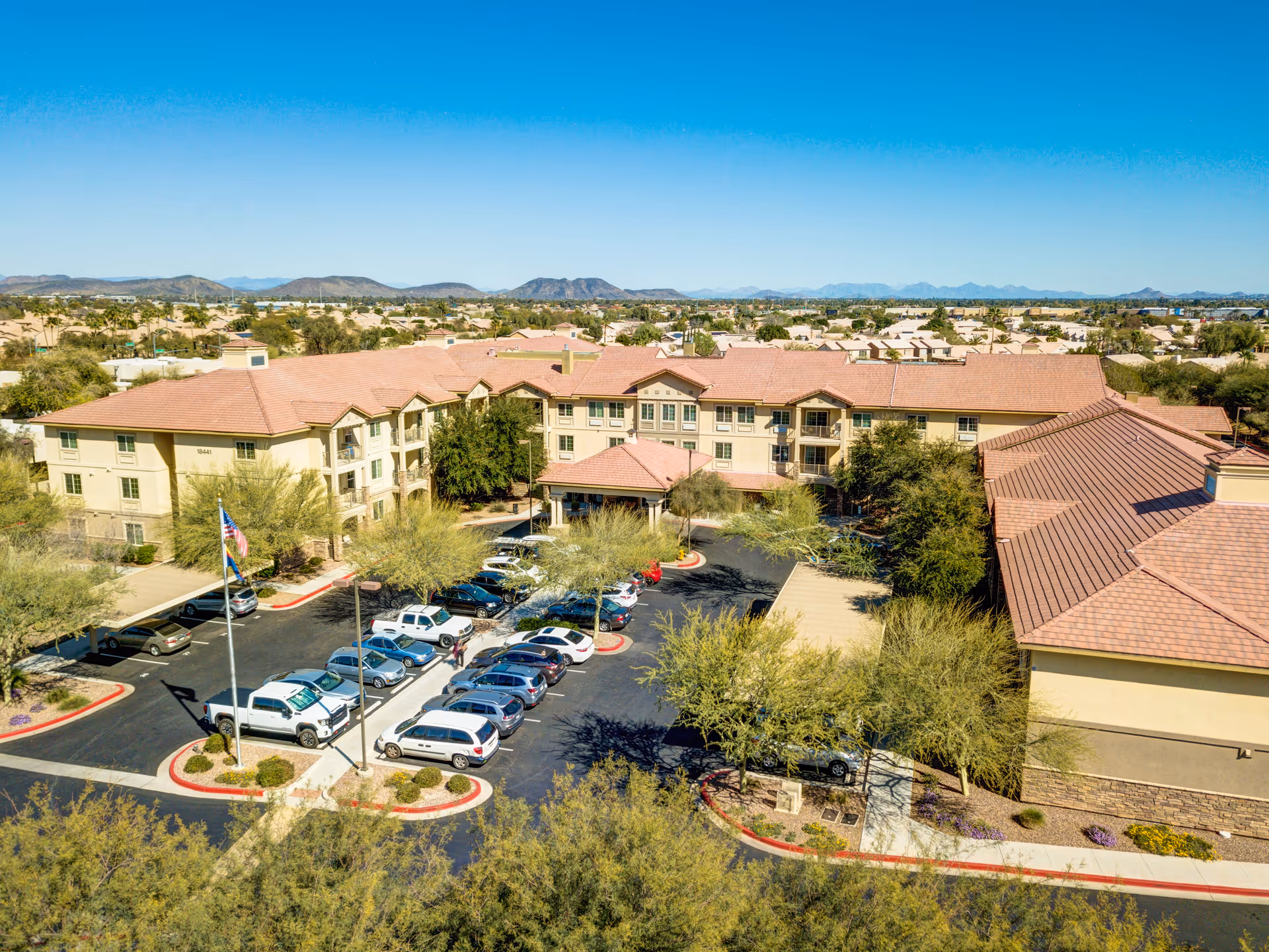 Aerial view of Palos Verdes Senior Living facility showing a large, multi-story building with a red-tiled roof surrounded by trees and a parking lot filled with cars. The background includes a suburban neighborhood and distant mountains under a clear blue sky.