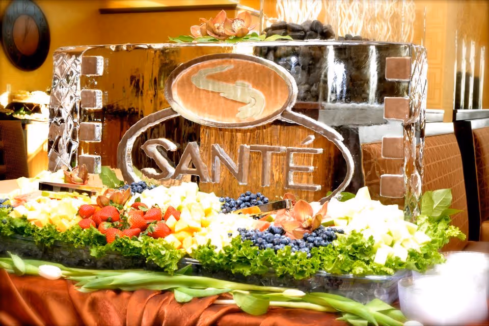 A decorative ice sculpture with the word 'SANTÉ' carved into it, surrounded by an assortment of fresh fruit including strawberries, blueberries, and pineapple, with green leafy garnish. The setting appears to be an indoor dining area with warm lighting and a clock on the wall in the background.