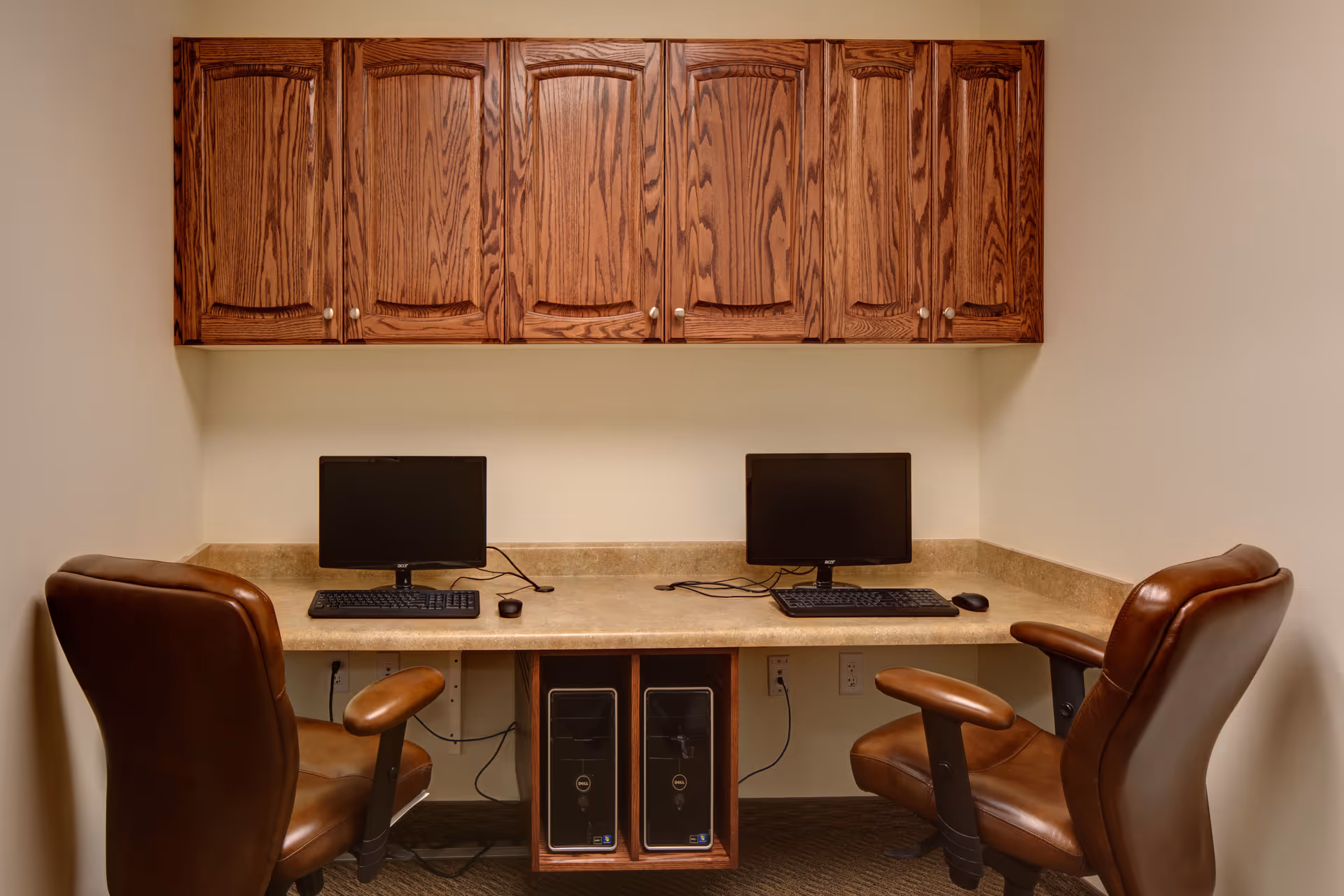 A small computer workstation area with two desktop computers, each with a monitor, keyboard, and mouse, placed on a beige countertop. Above the countertop are wooden cabinets with a natural finish. Two brown leather office chairs are positioned in front of the computers. The walls are light-colored and the floor is carpeted.