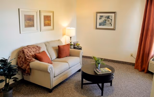 A cozy living room with a beige loveseat adorned with two rust-colored pillows and a matching throw blanket. A round black coffee table in front holds a small potted plant and some books. The room has beige carpet, light-colored walls with three framed artworks, a side table with a lamp, and rust-colored curtains partially covering a window with an air conditioning unit below.