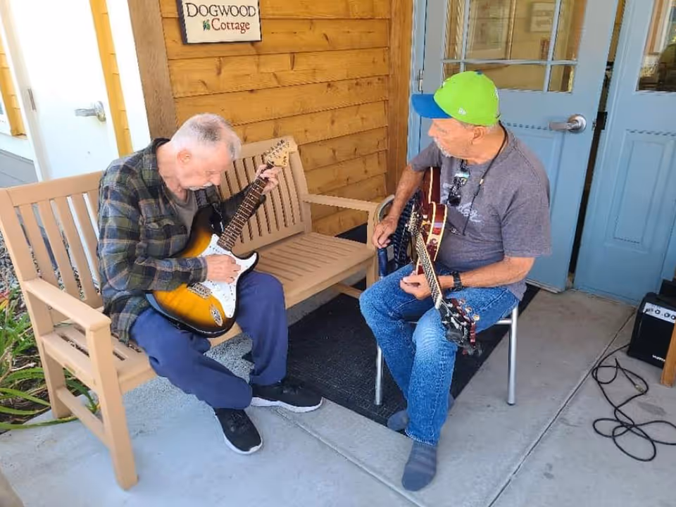 Two elderly men playing electric guitars outside a building with wooden siding and blue doors. One man is sitting on a wooden bench, and the other is sitting on a metal chair. There is a sign on the wall that reads 'DOGWOOD Cottage'.
