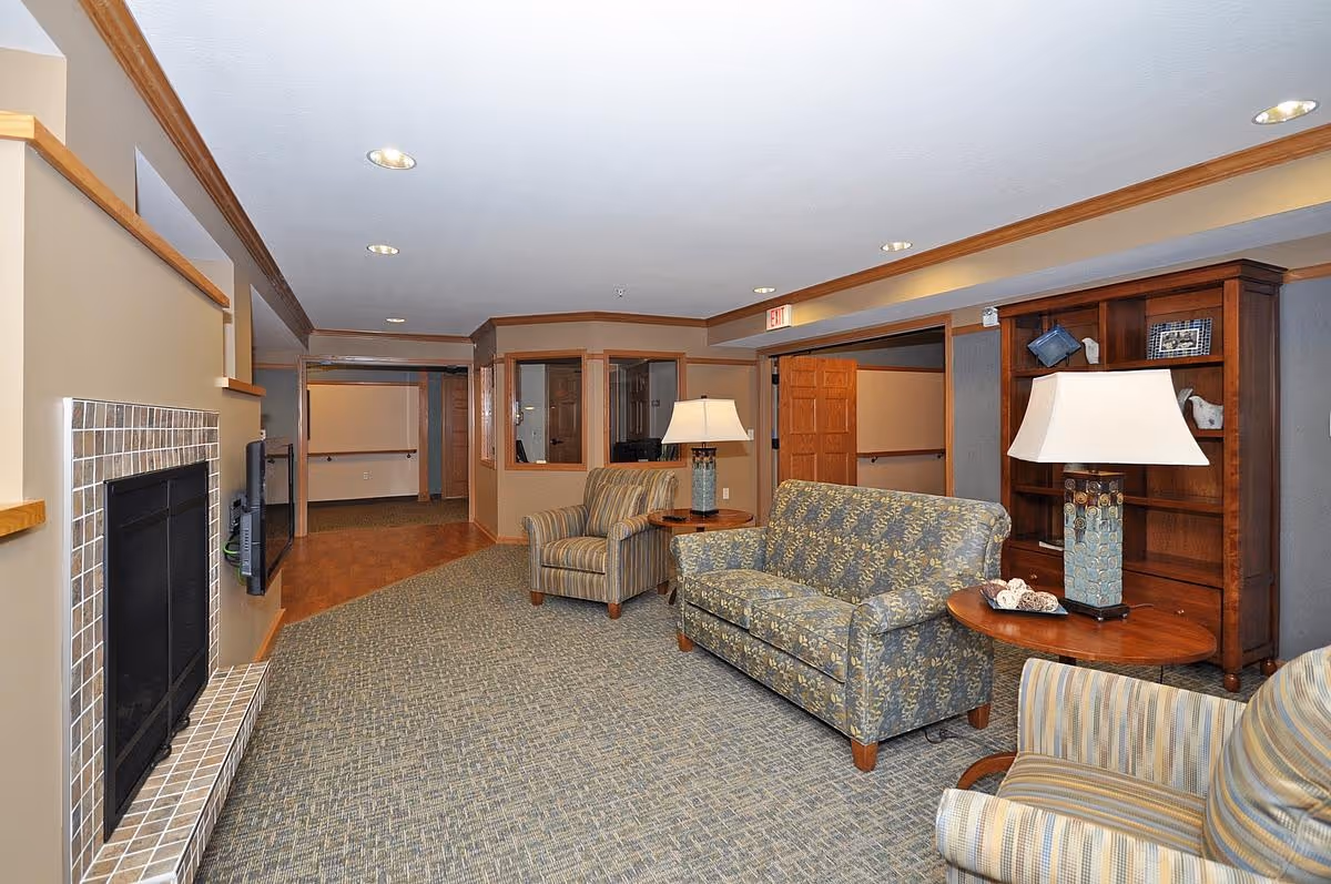 Comfortable common lounge with sofas, armchairs, lamps, shelving, and a fireplace in a senior living facility.