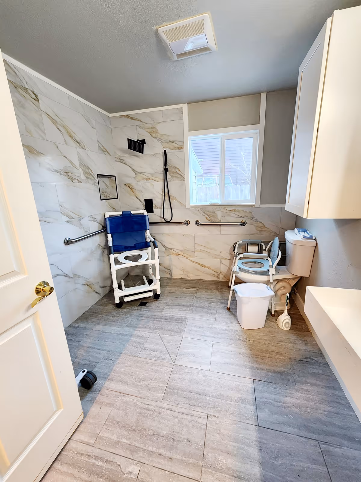 Accessible bathroom with a shower area featuring a blue shower chair, grab bars on the walls, a window, a toilet with a raised seat and armrests, a white trash bin, and a white cabinet above the toilet.