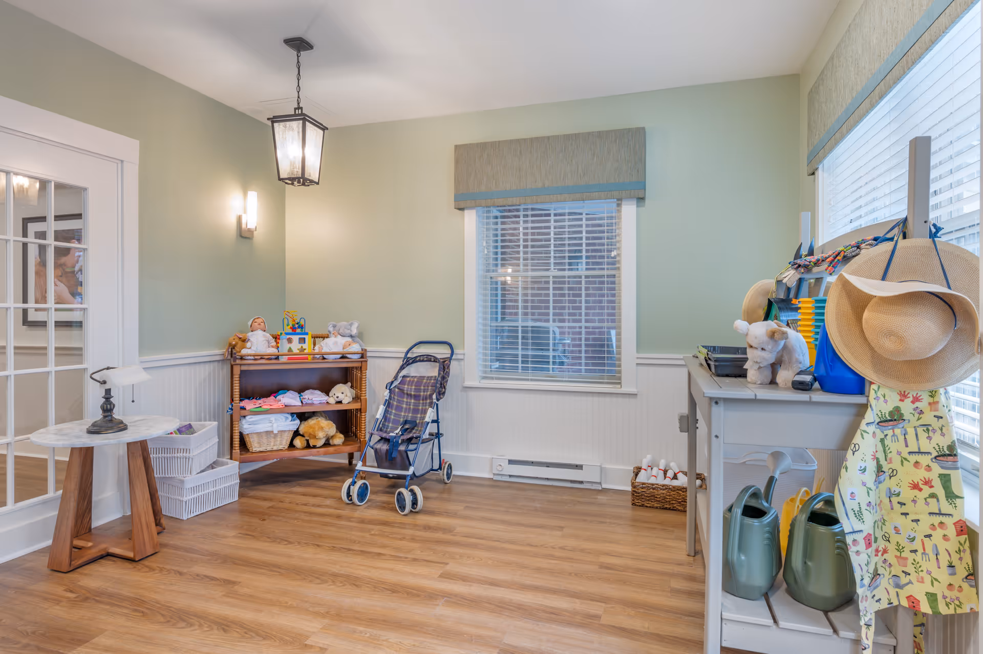 A bright room with light green walls and wood flooring featuring a small wooden shelf with dolls and stuffed animals, a baby stroller, a table with gardening tools including watering cans, a hat, and an apron hanging on a stand. There is a window with blinds and a valance, a small round table with a lamp, and a basket of toy bowling pins on the floor.