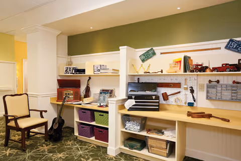 A cozy interior room with a green and yellow color scheme featuring a wooden chair with cream cushions, a black acoustic guitar resting against a white shelving unit, and a workbench area with various tools, books, and decorative items. The floor is carpeted with a green patterned design.