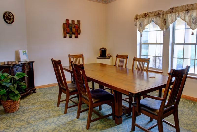 A dining room with a wooden rectangular table surrounded by six wooden chairs with dark cushions. There is a large window with floral valances letting in natural light. A small cabinet with a coffee maker is in the corner, and a decorative plant in a woven basket is on the floor next to a dark wooden sideboard. The walls are light-colored with minimal decorations including a clock and a wall art piece.