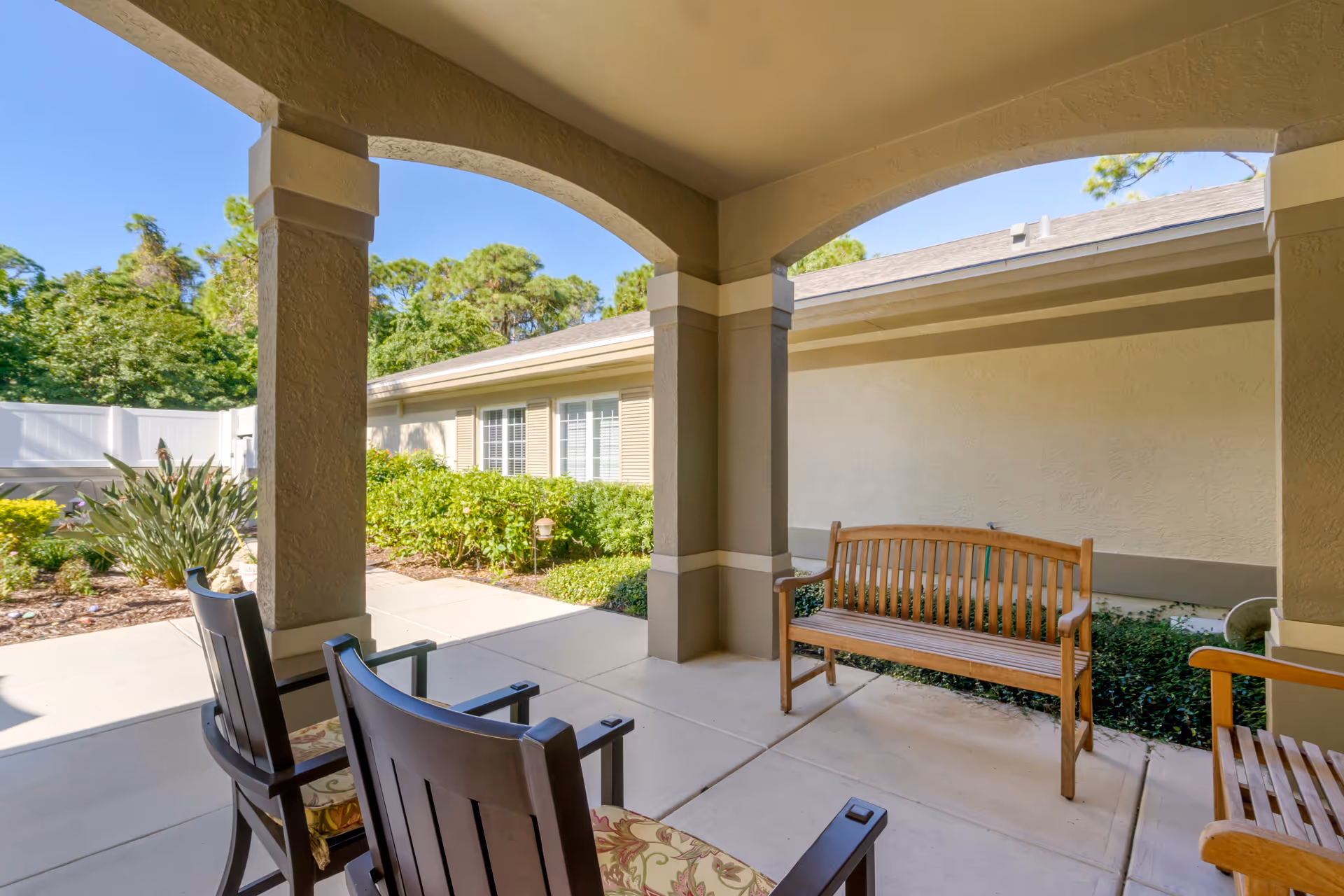 Covered outdoor patio area with two cushioned wooden chairs and a wooden bench, surrounded by beige walls and columns, with greenery and a clear blue sky in the background.
