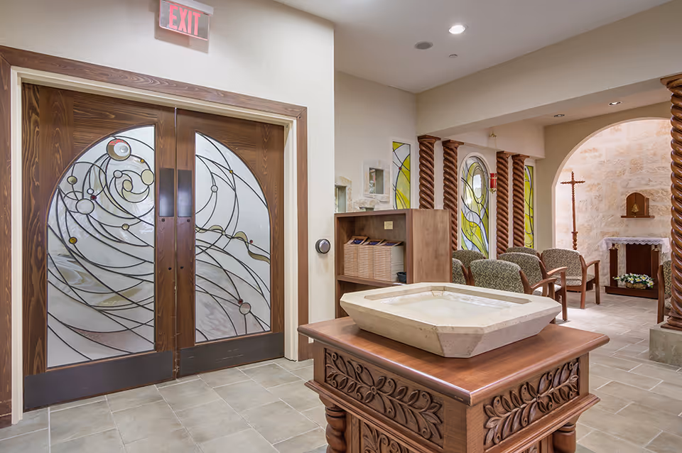 Interior view of a chapel or meditation room with decorative wooden double doors featuring stained glass designs, a wooden stand with a stone basin in the foreground, cushioned chairs arranged near stained glass windows, and a small altar with a cross and flowers in the background.