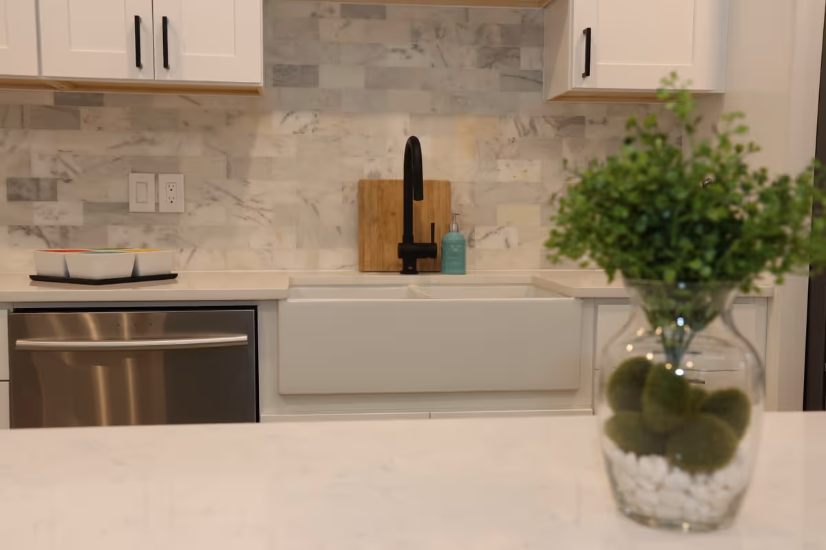 Modern kitchen area with a white farmhouse sink, black faucet, marble tile backsplash, white cabinets with black handles, a stainless steel dishwasher, and a countertop with a decorative glass vase containing green plants and moss balls.
