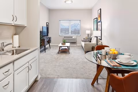 A bright and clean living area in an assisted living facility featuring a small kitchenette with a sink and white cabinets on the left, a round glass dining table set with plates, cups, and a bowl of lemons on the right, and a cozy living room with a beige sofa, coffee table, TV on a stand, and a window with blinds in the background.