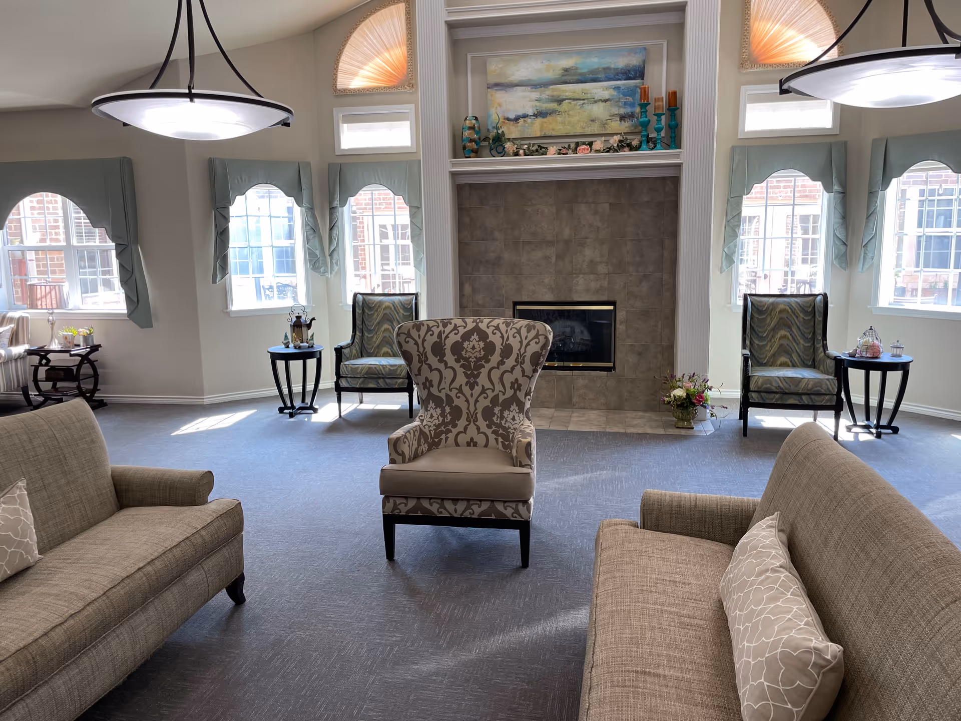 A bright and spacious living room with two beige sofas facing each other and a patterned armchair in the center. The room features a fireplace with a large painting above it, flanked by two windows with light blue curtains. Two side tables with decorative items are placed near the windows, and two modern ceiling lights hang from the ceiling.