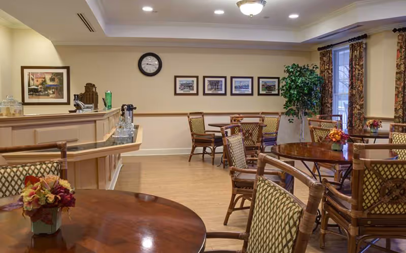 A cozy dining area with several round wooden tables and cushioned chairs arranged around them. The room has beige walls adorned with framed pictures and a clock. There is a counter on the left side with a coffee maker and glass containers. A large window with patterned curtains lets in natural light, and a potted plant is placed near the window.