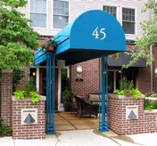 Entrance to a senior living facility with a blue canopy displaying the number 45, flanked by brick pillars with decorative planters and greenery around the doorway.