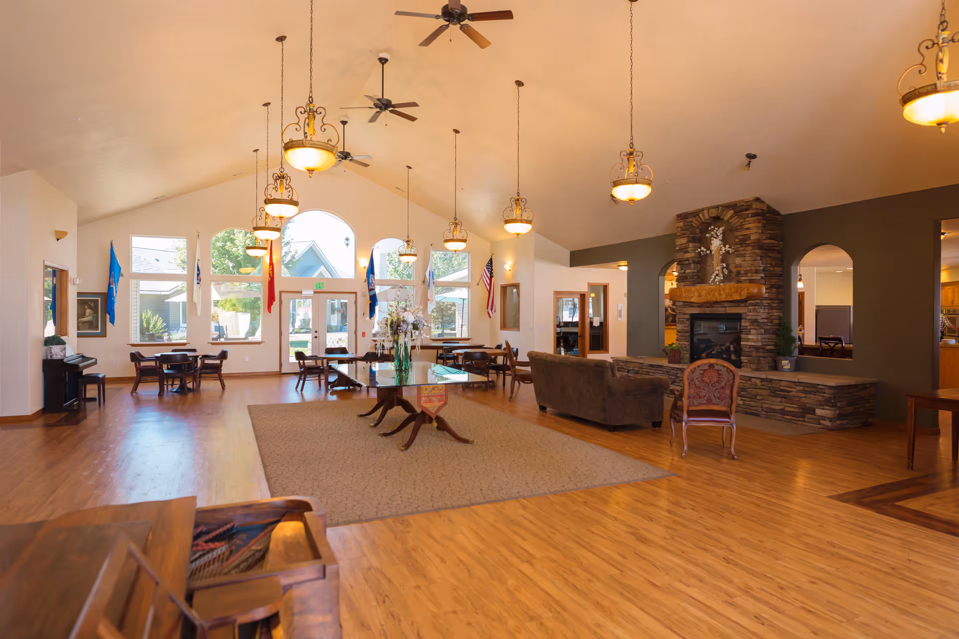 Spacious common area in a senior living facility with wooden floors, a large carpeted area, multiple tables and chairs near windows with flags hanging above, a stone fireplace with a mantel, ceiling fans, and hanging light fixtures.