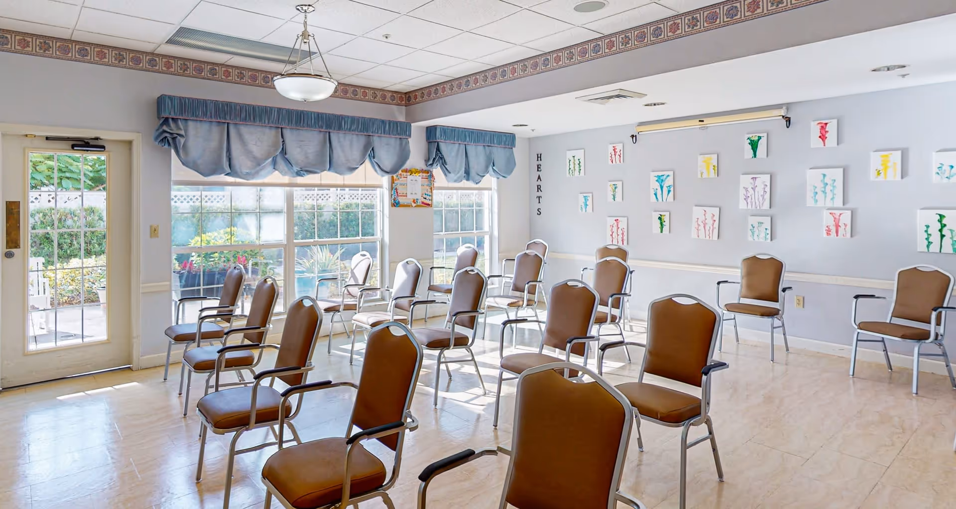 A bright room with multiple brown cushioned chairs arranged in rows facing the same direction. Large windows with blue valances let in natural light, and there is a door leading outside. The walls are decorated with small colorful artwork and the word 'HEARTS' is displayed vertically on one wall.