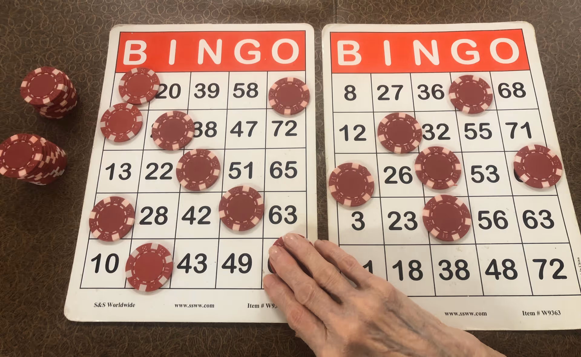 Two bingo cards on a table with red chip markers and a hand covering a number.