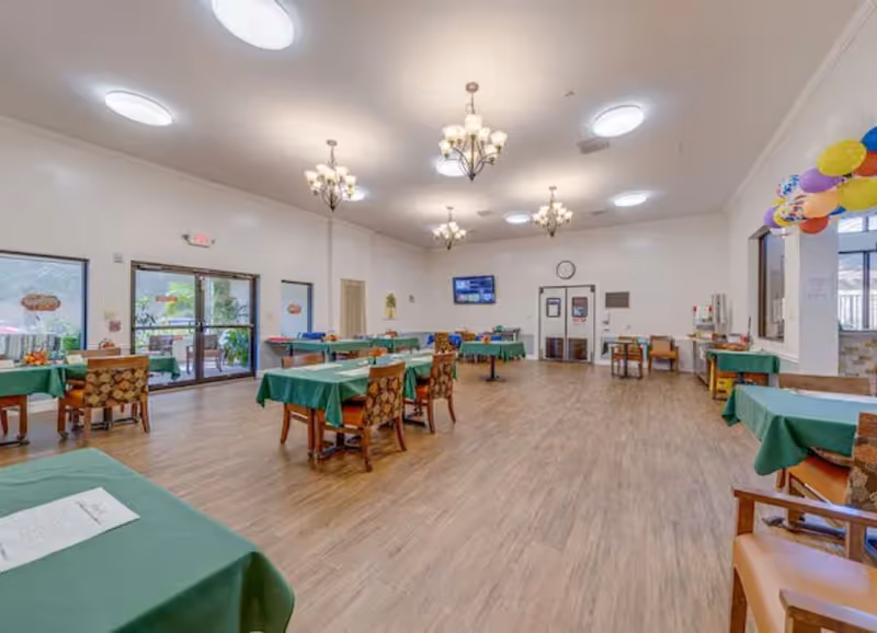 A spacious dining room with several tables covered in green tablecloths and chairs arranged around them. The room has wooden flooring, white walls, and multiple chandeliers hanging from the ceiling. There are large windows and glass doors on one side, allowing natural light to enter. Colorful balloons are visible on the right side near a window. A television and clock are mounted on the far wall.