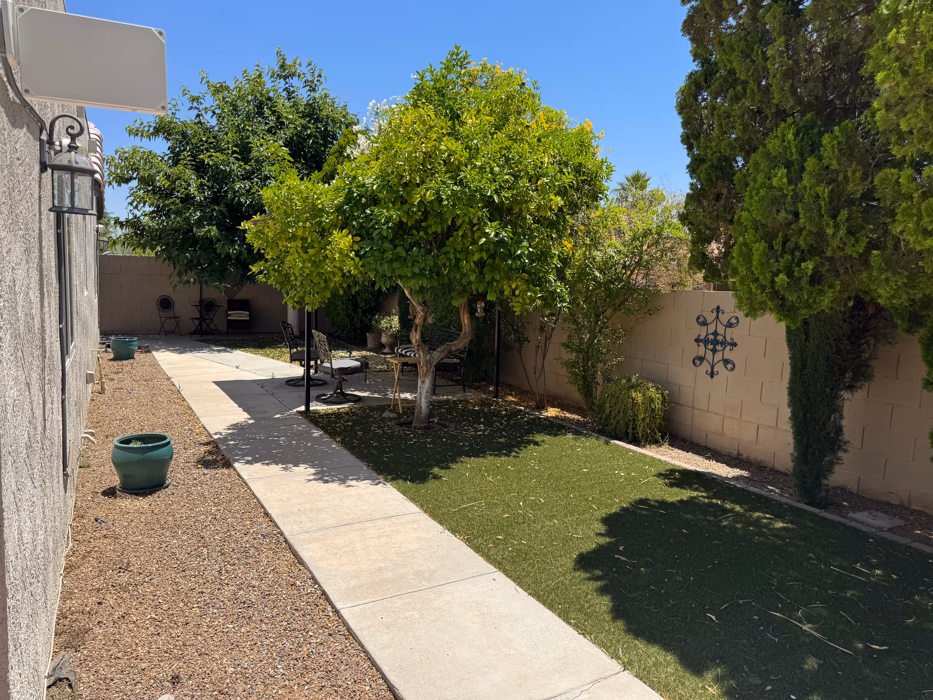 Outdoor garden area with a concrete walkway, green trees, shrubs, and a small seating area with chairs and a table. The area is enclosed by a beige brick wall and has a clear blue sky above.