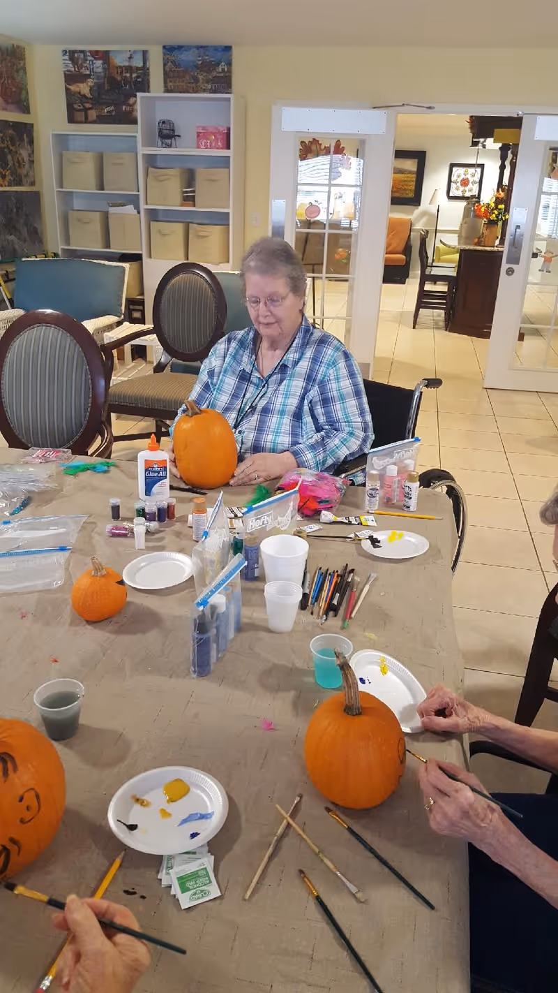 An elderly woman sitting at a table in a wheelchair, painting a pumpkin. The table is covered with art supplies including paintbrushes, paint bottles, glue, and paper plates with paint. Other pumpkins and hands of other participants are visible, indicating a group pumpkin painting activity in a well-lit room with chairs and shelves in the background.
