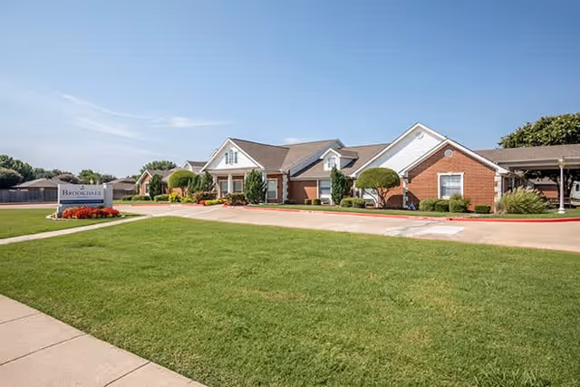 Exterior view of Brookdale Watauga showing the building entrance, landscaped lawn, and clear blue sky.