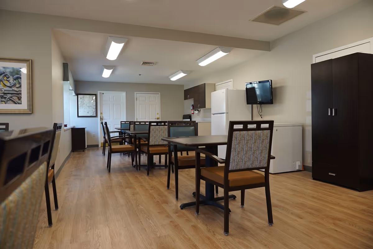Interior view of a communal dining area in a senior living facility with multiple tables and chairs arranged on a wooden floor. The room features a refrigerator, a small wall-mounted TV, a dark wooden cabinet, and fluorescent ceiling lights. There are two white doors at the far end and framed artwork on the walls.