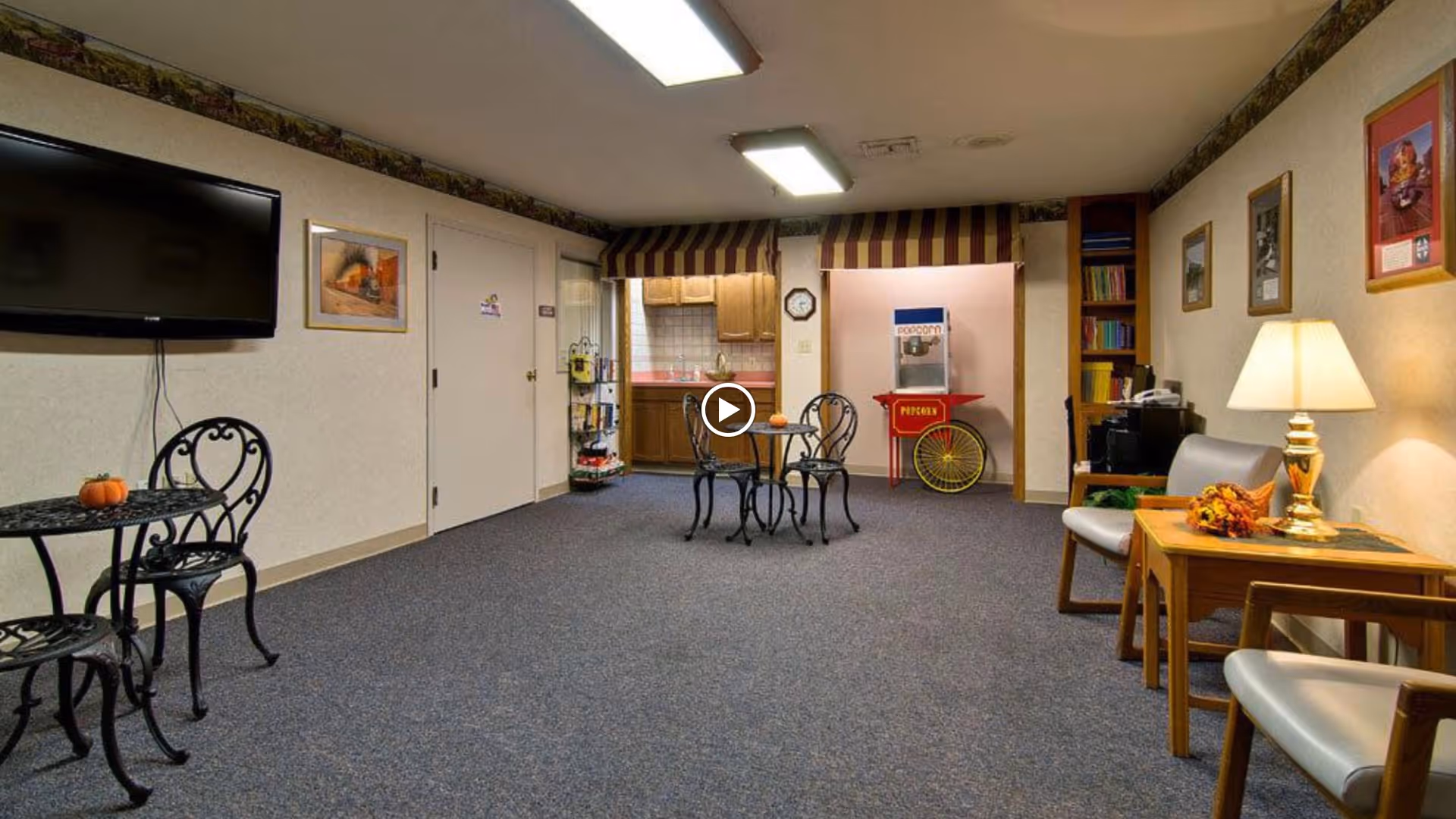 A cozy common area with carpeted floor featuring a small round table with two wrought iron chairs, a wall-mounted flat screen TV, and a kitchenette in the background. There is a popcorn machine on a red cart, a bookshelf, and a seating area with two chairs and a wooden side table with a lamp and autumn decorations. The walls are decorated with framed pictures.