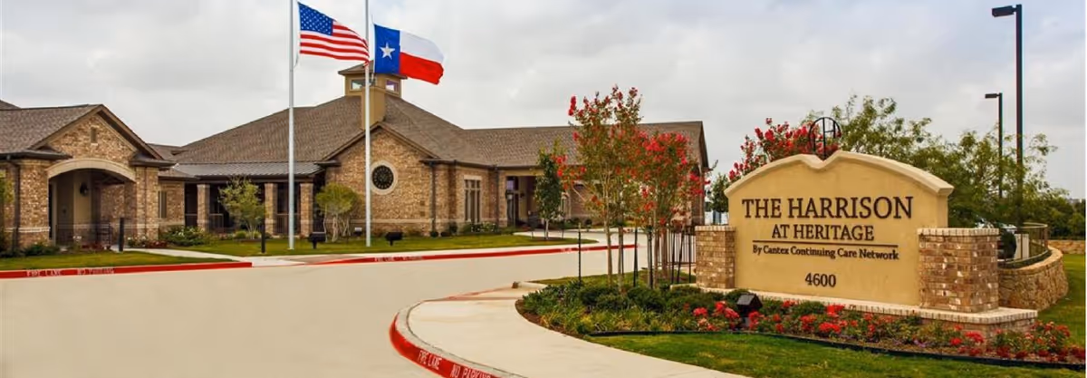 Exterior view of The Harrison At Heritage senior living facility showing a brick building with a peaked roof, two flagpoles with the American and Texas flags, a landscaped area with trees and flowers, and a large sign with the facility name and address.