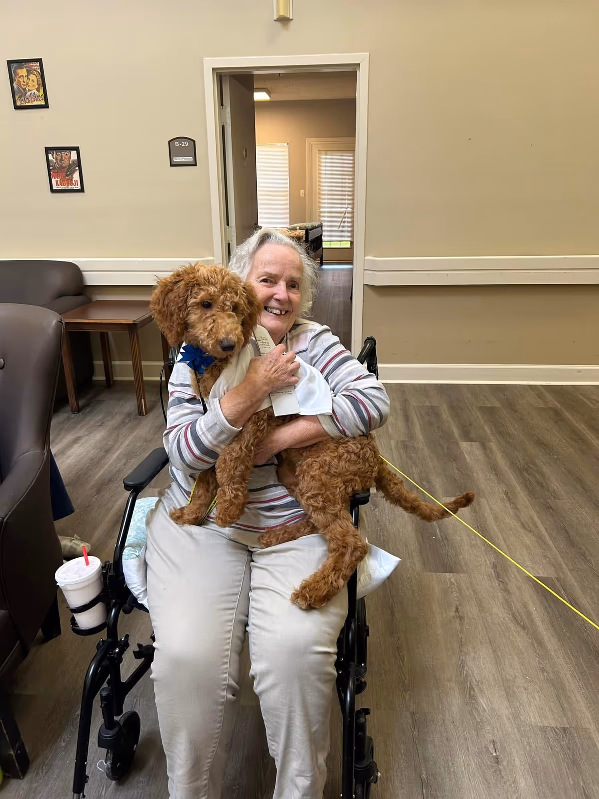 An elderly woman sitting in a wheelchair inside a senior living facility, smiling and holding a curly-haired brown dog wearing a white shirt and blue collar. The room has wooden flooring, beige walls, and some framed pictures on the wall.