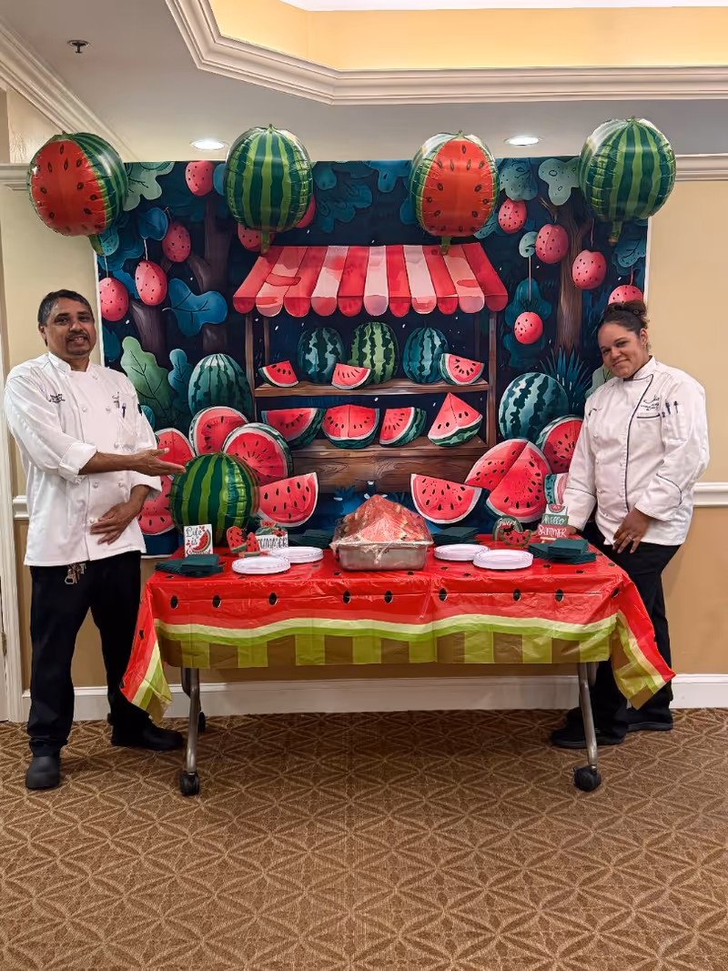 Two chefs standing on either side of a table decorated with a watermelon-themed tablecloth and watermelon balloons. Behind them is a colorful backdrop featuring illustrations of watermelons and a fruit stand. The table has plates, napkins, and a tray with watermelon slices.