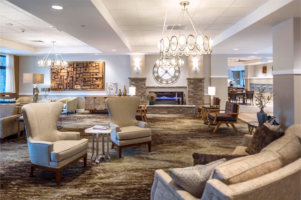 A spacious and well-lit senior living facility common area with comfortable armchairs and sofas arranged around small tables. The room features a large decorative wall clock above a stone fireplace, modern chandeliers, and artistic wall decor. The space has a warm and inviting atmosphere with neutral tones and soft lighting.