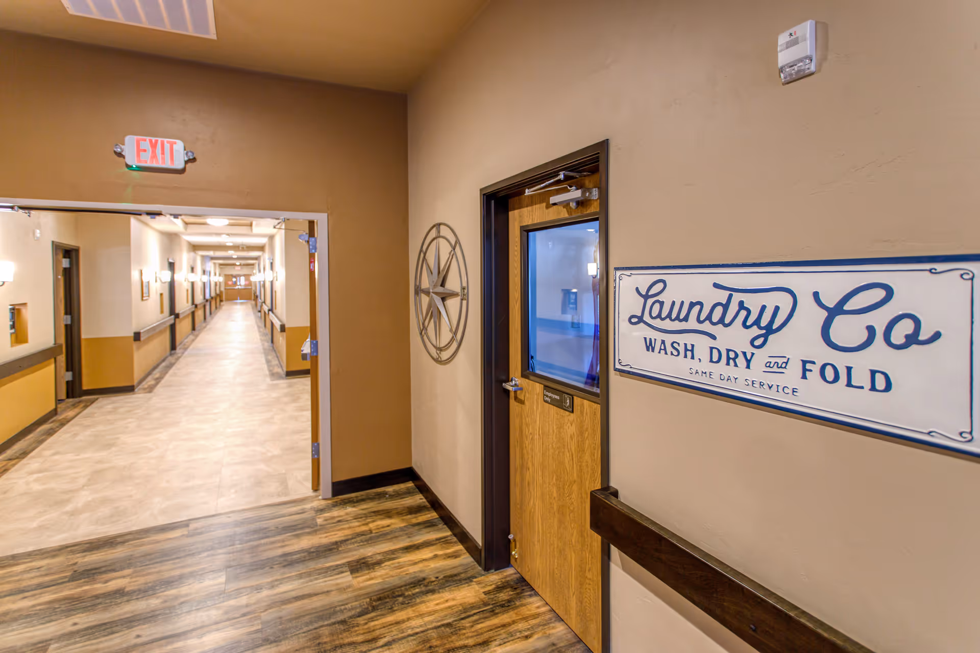 Interior hallway of a senior living facility with beige walls and wood-patterned flooring. A door labeled 'Laundry Co Wash, Dry and Fold Same Day Service' is visible on the right side. The hallway extends into the distance with multiple doors and wall-mounted lights. An exit sign is above the open doorway.