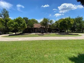 A single-story brick building with a brown roof surrounded by green trees and a well-maintained lawn under a blue sky with some clouds. A paved walkway curves in front of the building, and there is a sign in front of the building.