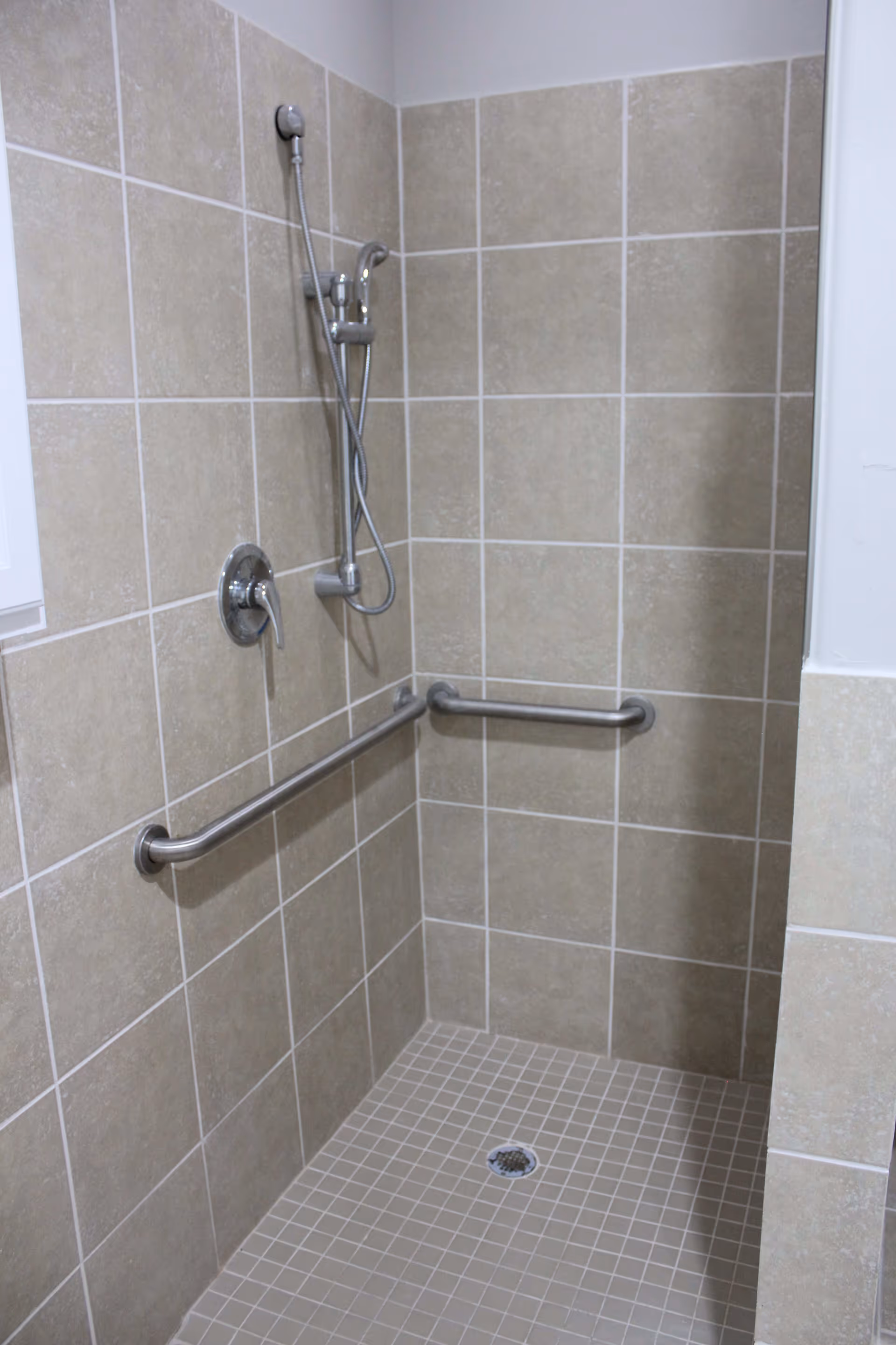 A tiled walk-in shower with beige tiles on the walls and floor, equipped with a handheld showerhead and two stainless steel grab bars for support.