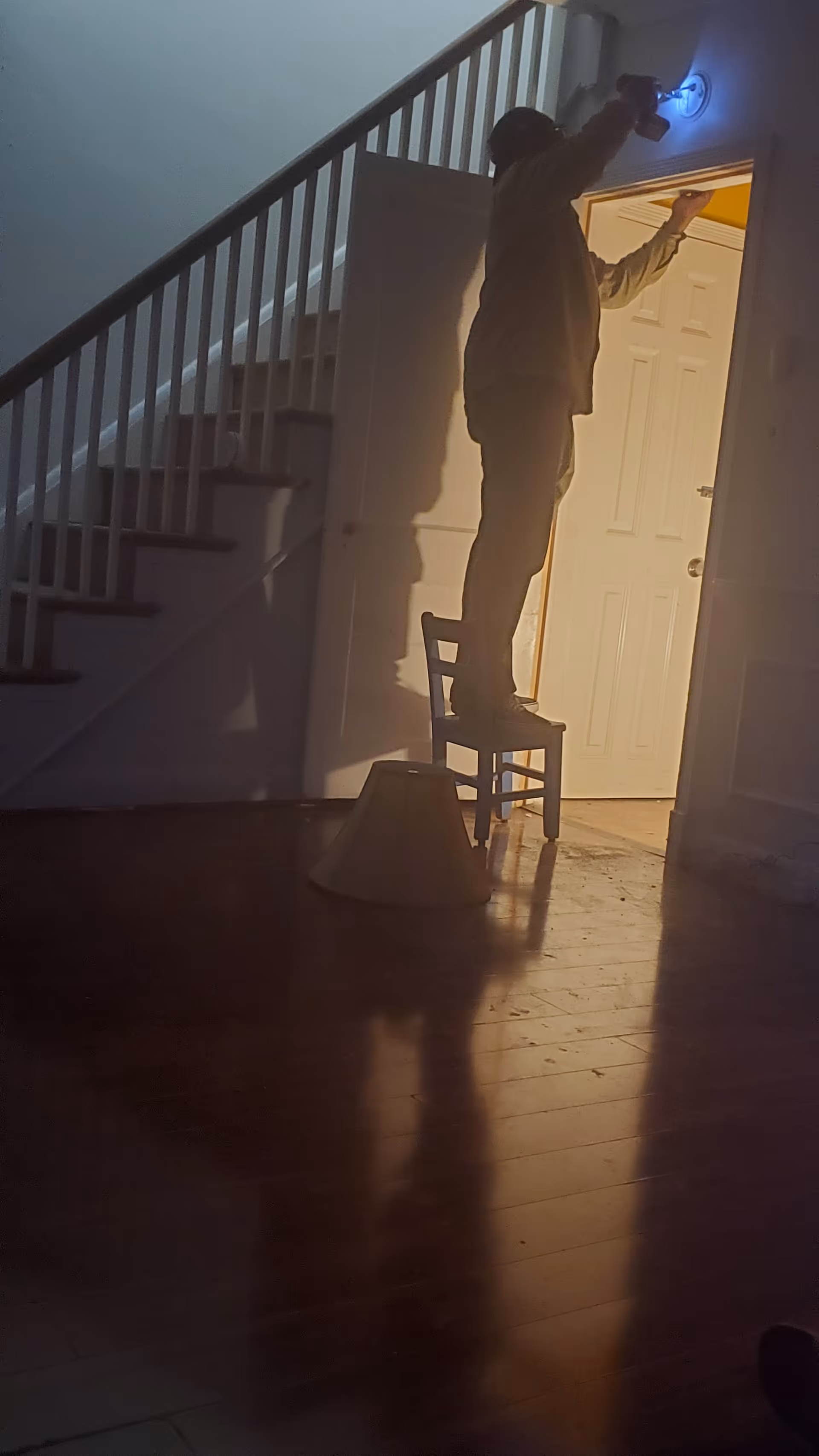 A person standing on a chair in a dimly lit room, reaching up to adjust or install a small blue light fixture near the ceiling. The room has wooden floors, a staircase with white railings, and an open door leading to a brightly lit area.