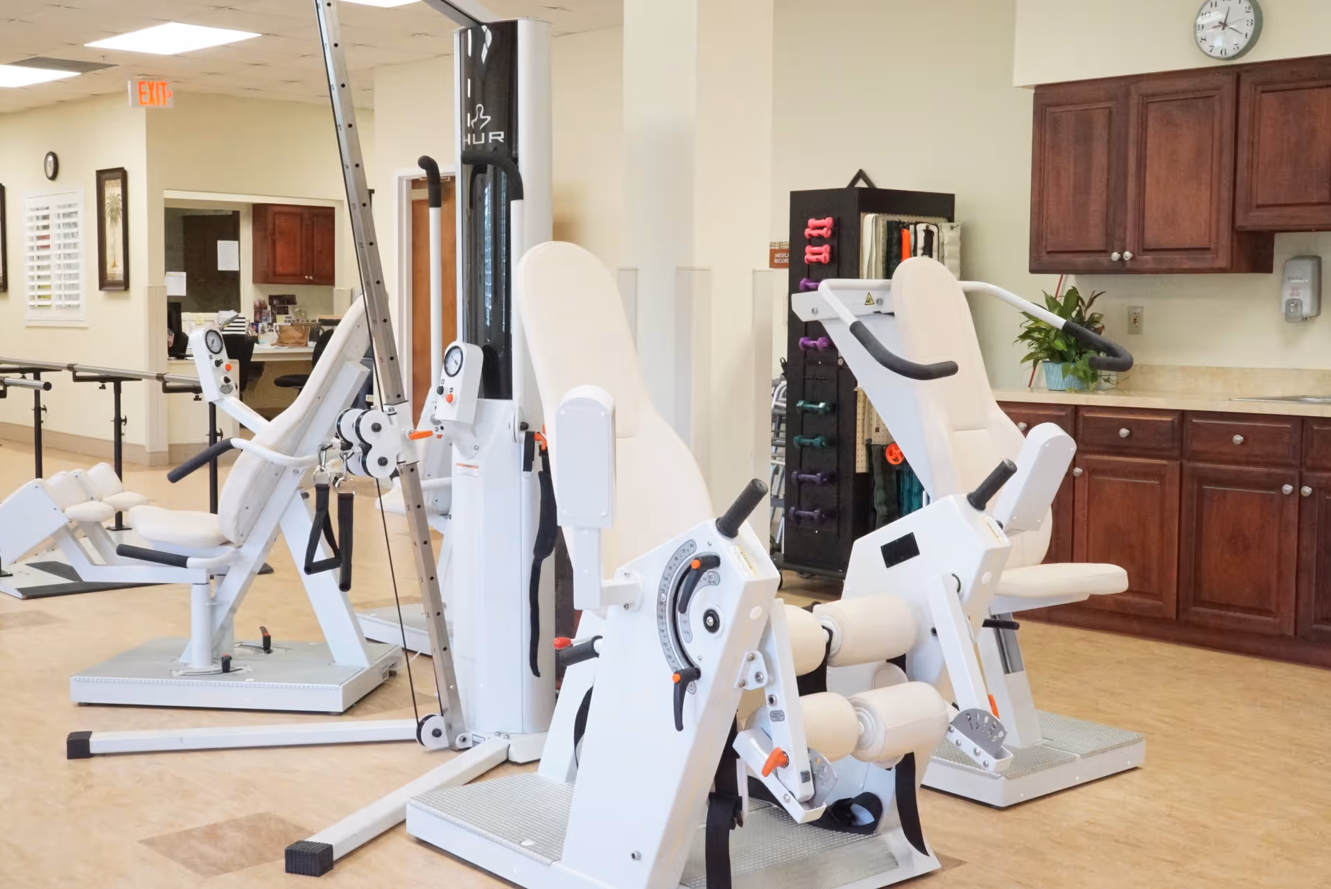 A fitness room in a senior living facility featuring white exercise machines designed for seated workouts. The room has wooden cabinets, a clock on the wall, and a rack with colorful dumbbells and exercise bands. The floor is light-colored wood, and the space is well-lit with overhead lighting.