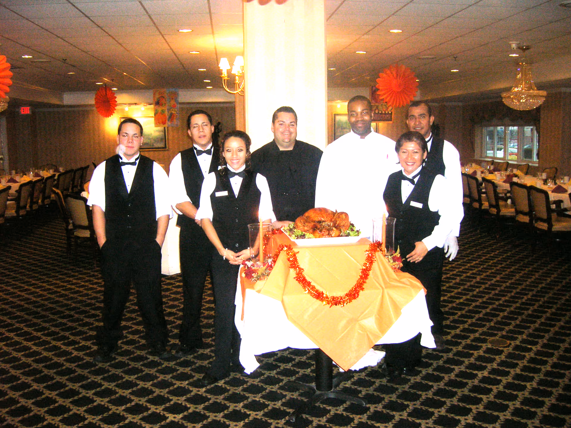 A group of catering staff in formal uniforms pose behind a decorated table with a roasted turkey in a large dining room.