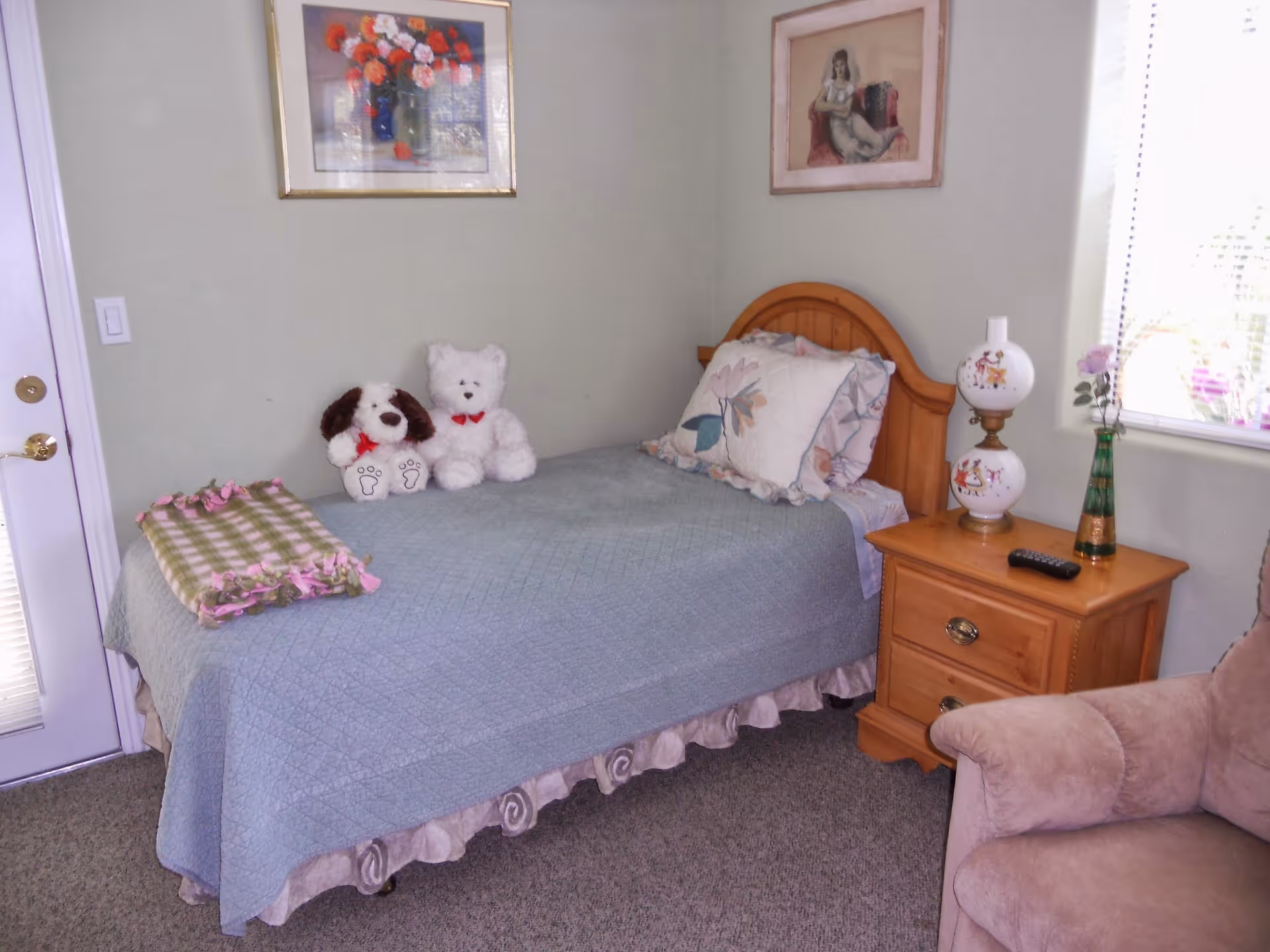 A cozy bedroom with a single wooden bed covered with a light blue quilt and floral pillows. Two stuffed animals, a dog and a bear, sit on the bed. A wooden nightstand next to the bed holds a decorative lamp, a remote control, and a green vase with flowers. There are two framed pictures on the light-colored walls, and a window with blinds lets in natural light. A soft armchair is partially visible in the foreground.