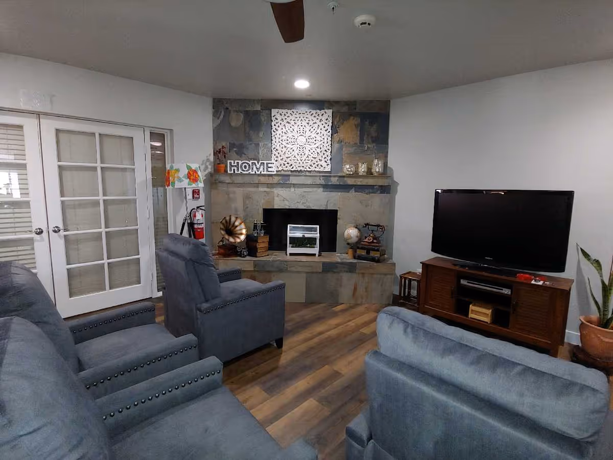 A cozy living room with gray upholstered chairs and sofas arranged around a stone fireplace. The fireplace mantel is decorated with a white decorative panel, a 'HOME' sign, and various small decorative items. To the right, there is a wooden TV stand with a flat-screen television and a potted plant beside it. French doors with glass panes are visible on the left side of the room.