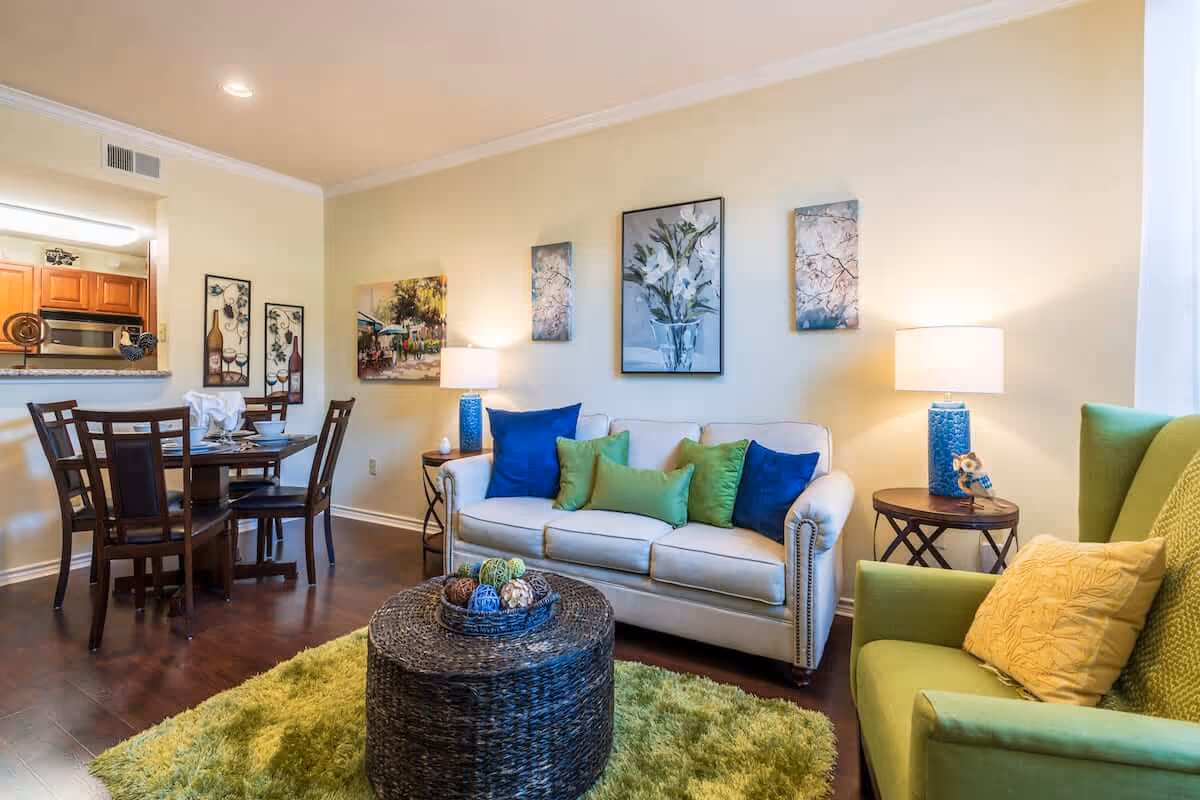 Bright living room with a white sofa, green armchair, colorful pillows, a round wicker coffee table on a green rug, and a dining area visible in the background.