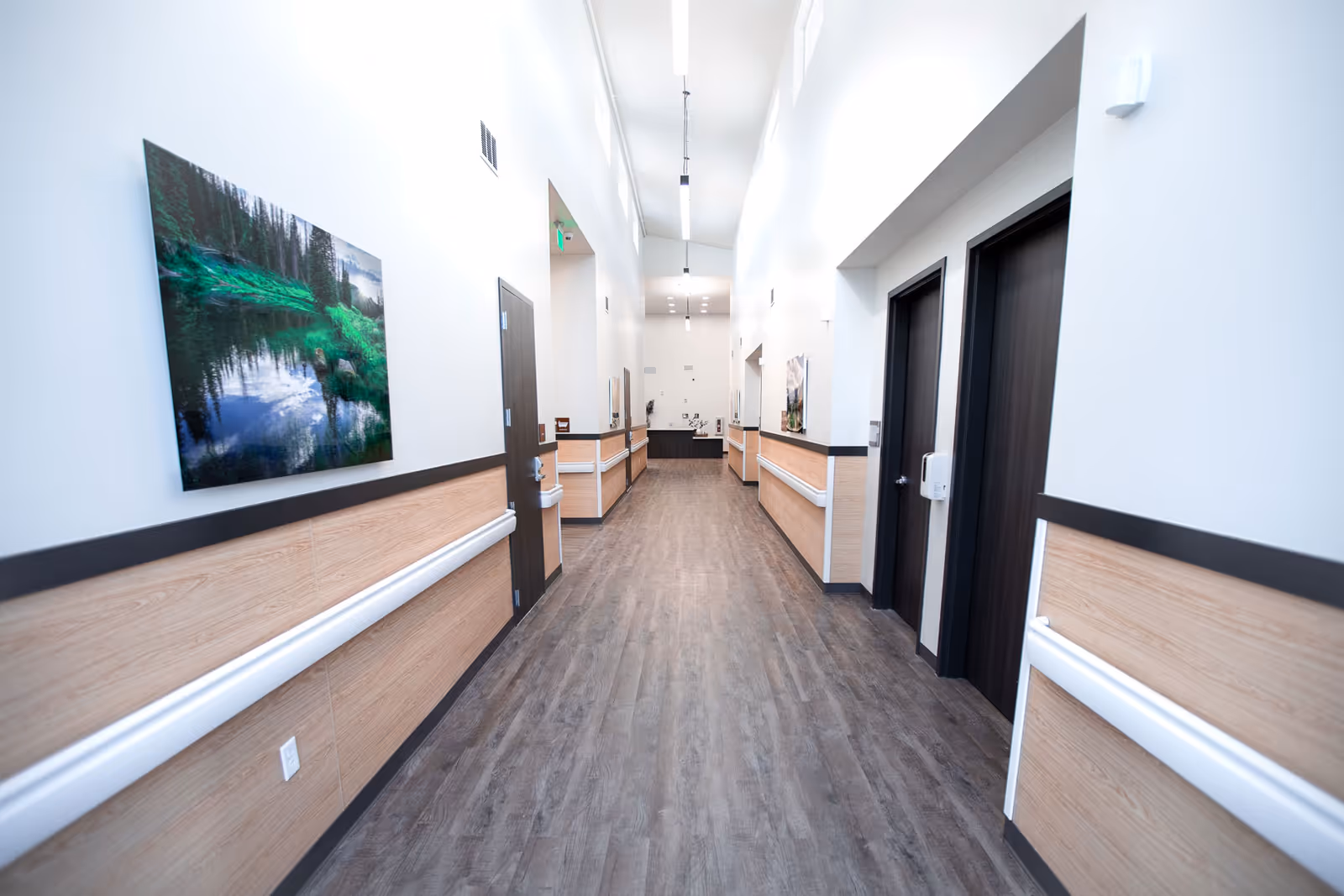 A clean, well-lit hallway in a senior living facility with wood-patterned flooring and white walls featuring wooden paneling halfway up. The hallway has several dark wooden doors on both sides, handrails along the walls, and landscape artwork hanging on the left wall. Bright ceiling lights illuminate the corridor.