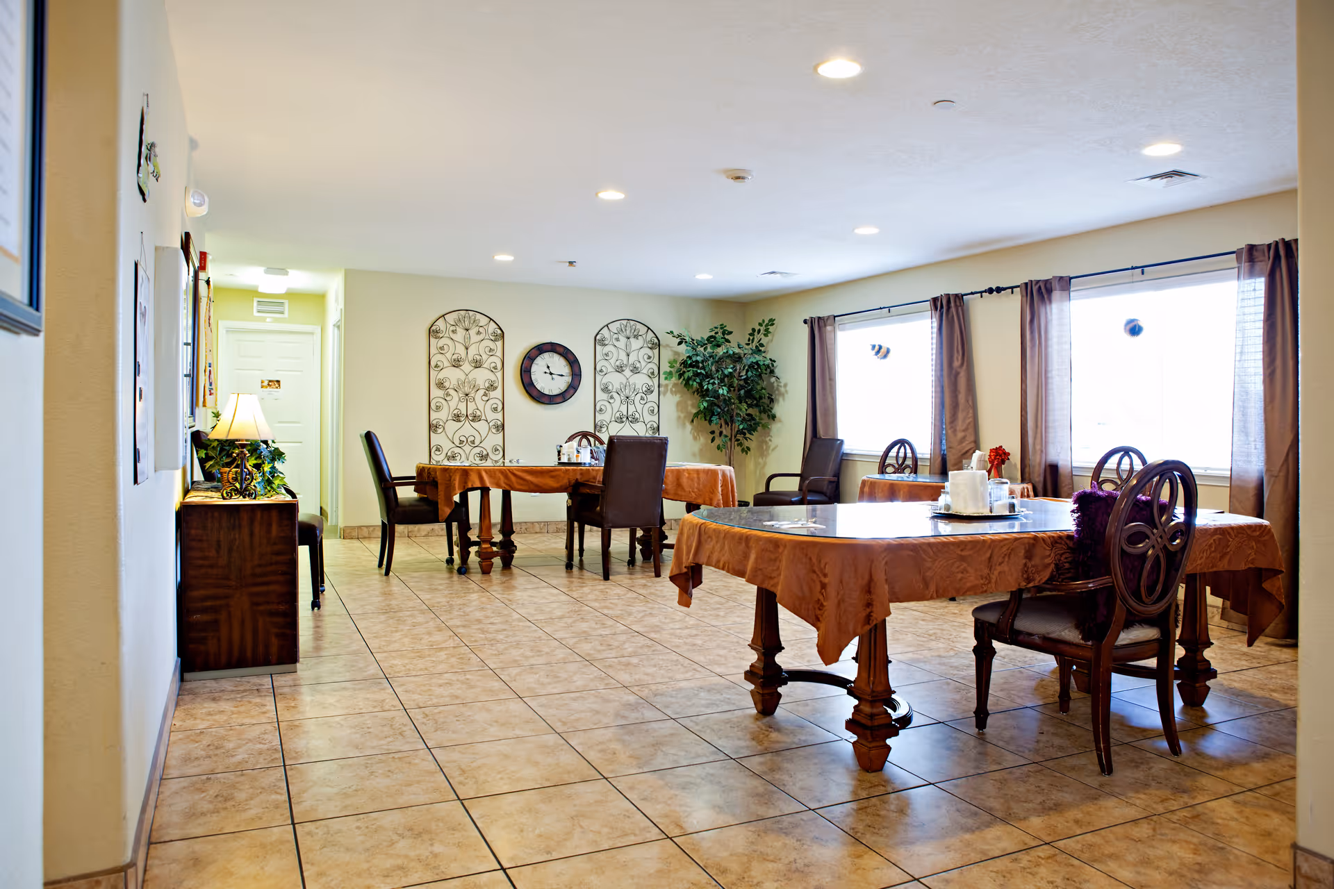 A spacious dining room with tiled floors and several wooden tables covered with brown tablecloths. Each table is surrounded by wooden chairs with cushioned seats. The room has large windows with brown curtains allowing natural light to fill the space. Decorative wall art and a clock are mounted on the far wall, and a potted plant is placed in the corner. A small wooden cabinet with a lamp and some greenery is visible on the left side near the entrance.