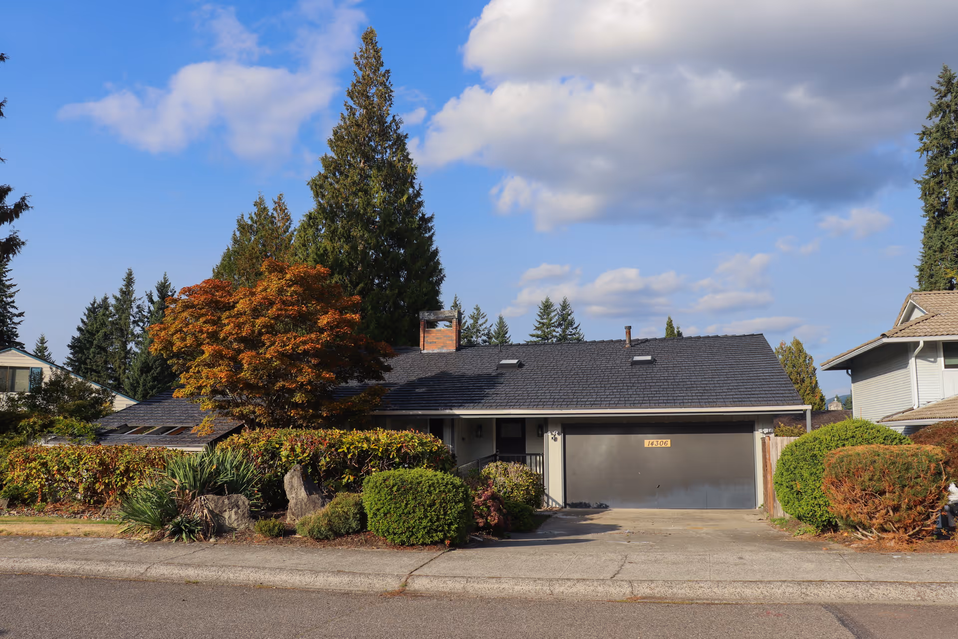 Front exterior view of a single-story house with a dark roof, a large garage door, and a variety of trees and shrubs in the front yard under a partly cloudy sky.