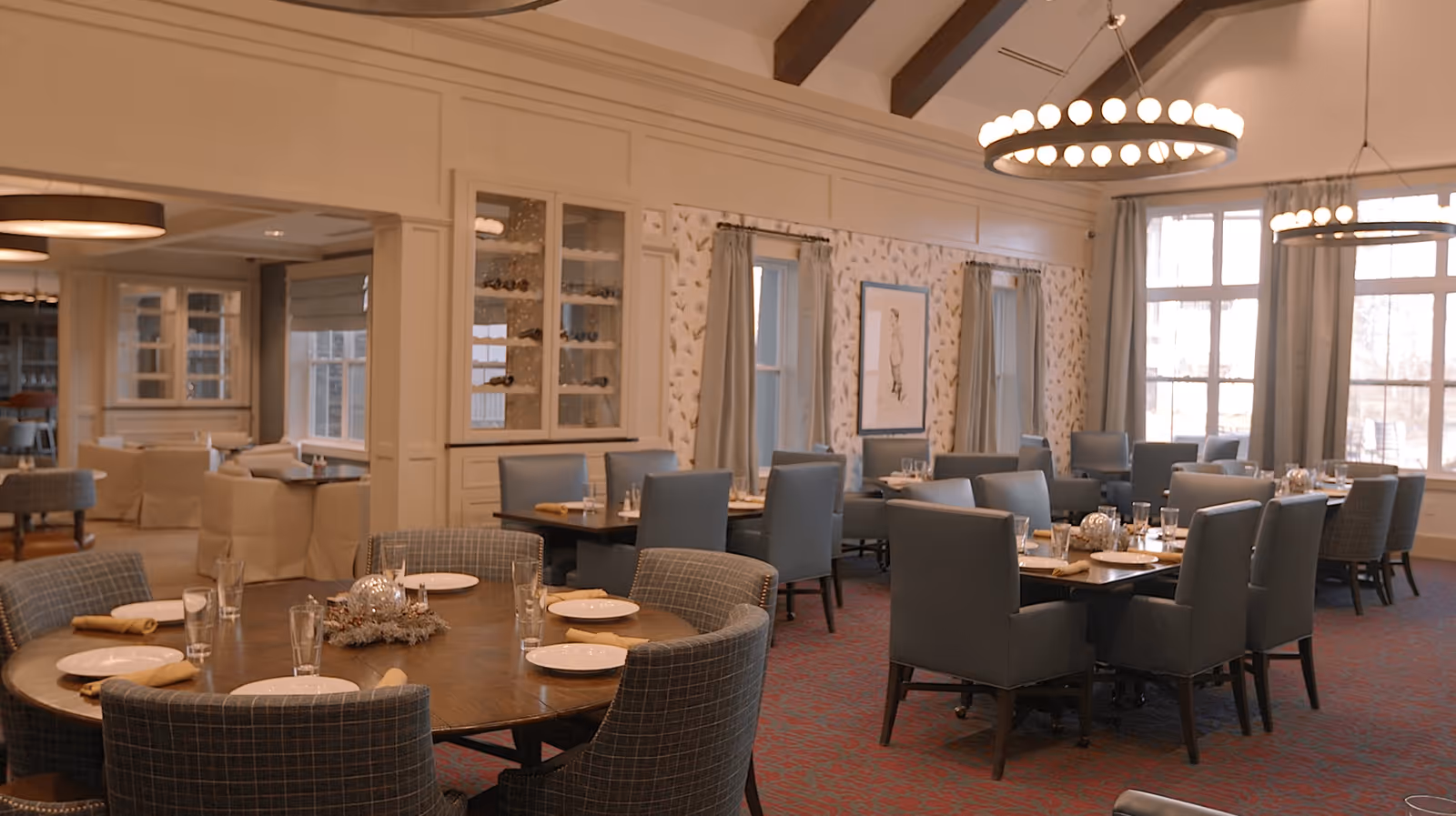 A spacious dining room with multiple tables set with plates, glasses, and napkins. The room features large windows with curtains, patterned wallpaper, and modern circular chandeliers hanging from a ceiling with exposed wooden beams. Upholstered chairs surround the tables, and a cabinet with glass doors displaying wine bottles is visible.