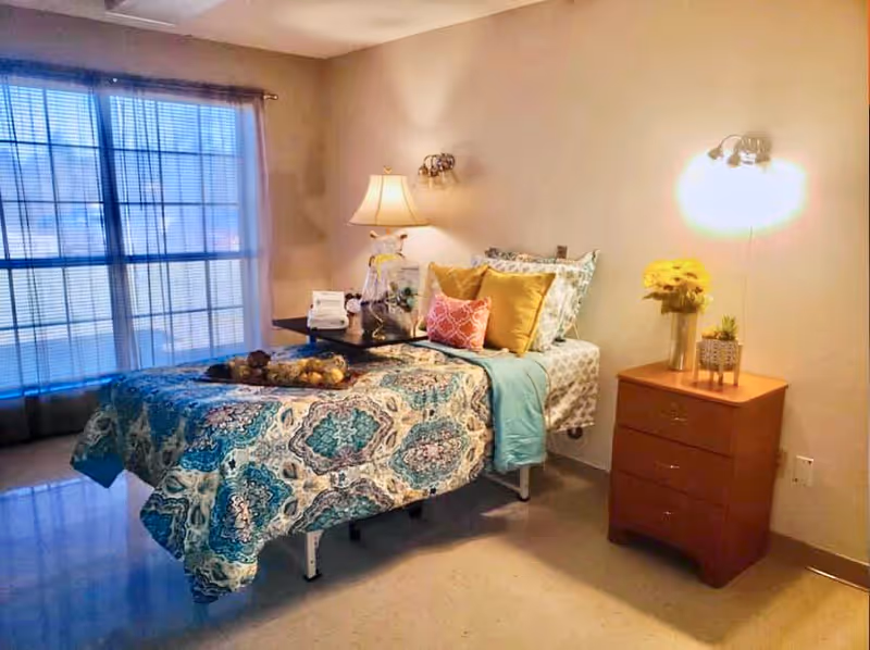 A neatly made single bed with a patterned blue and white bedspread, multiple pillows in yellow and pink, and a teal throw blanket. The bed has a small overbed table with decorative items on it. Next to the bed is a wooden nightstand with a vase of yellow flowers and a small plant. The room has a large window with sheer curtains allowing natural light to enter. Two wall-mounted lights and a table lamp provide additional lighting.