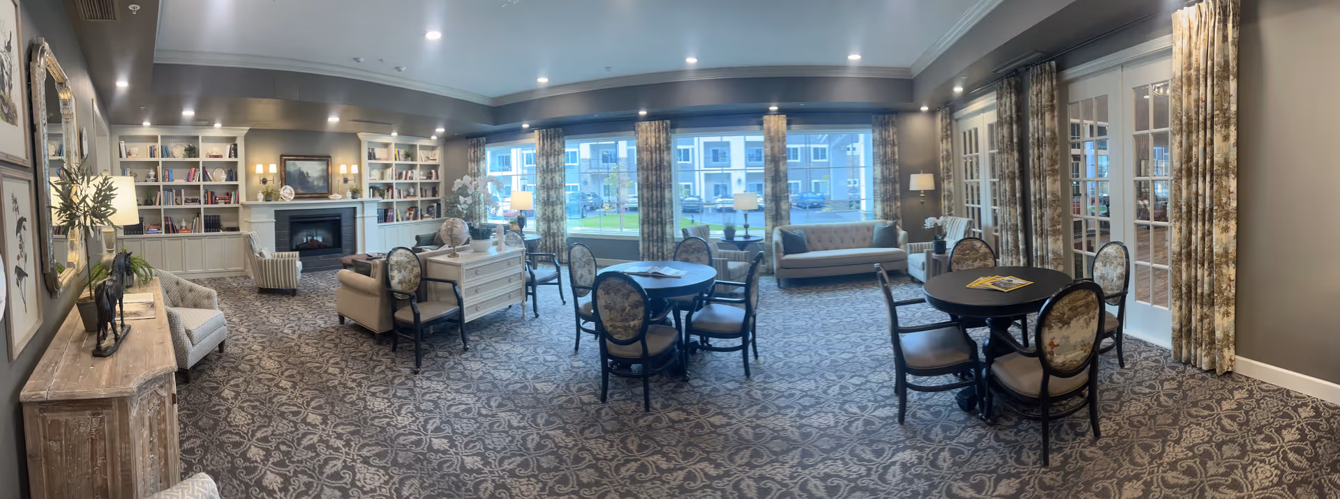 A spacious and elegantly decorated living room in an assisted living community featuring patterned carpet, multiple round tables with chairs, comfortable sofas and armchairs, large windows with floral curtains allowing natural light, built-in bookshelves, a fireplace, and decorative lamps and artwork.